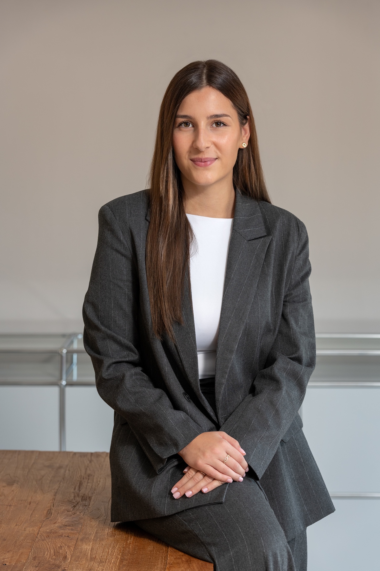 Portrait einer Frau mit langen braunen Haaren in grauem Anzug mit feinen Streifen, sitzend auf einem Holztisch.
