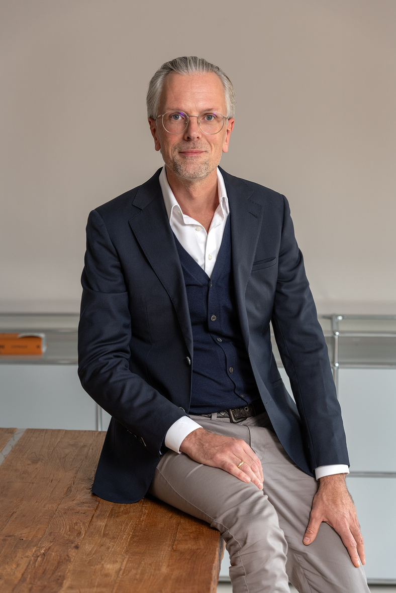 Mann mit Brille, grauen Haaren und Bart trägt weißen Hemd, dunkelblauen Anzug und sitzt auf einem Holztisch vor grauer Wand