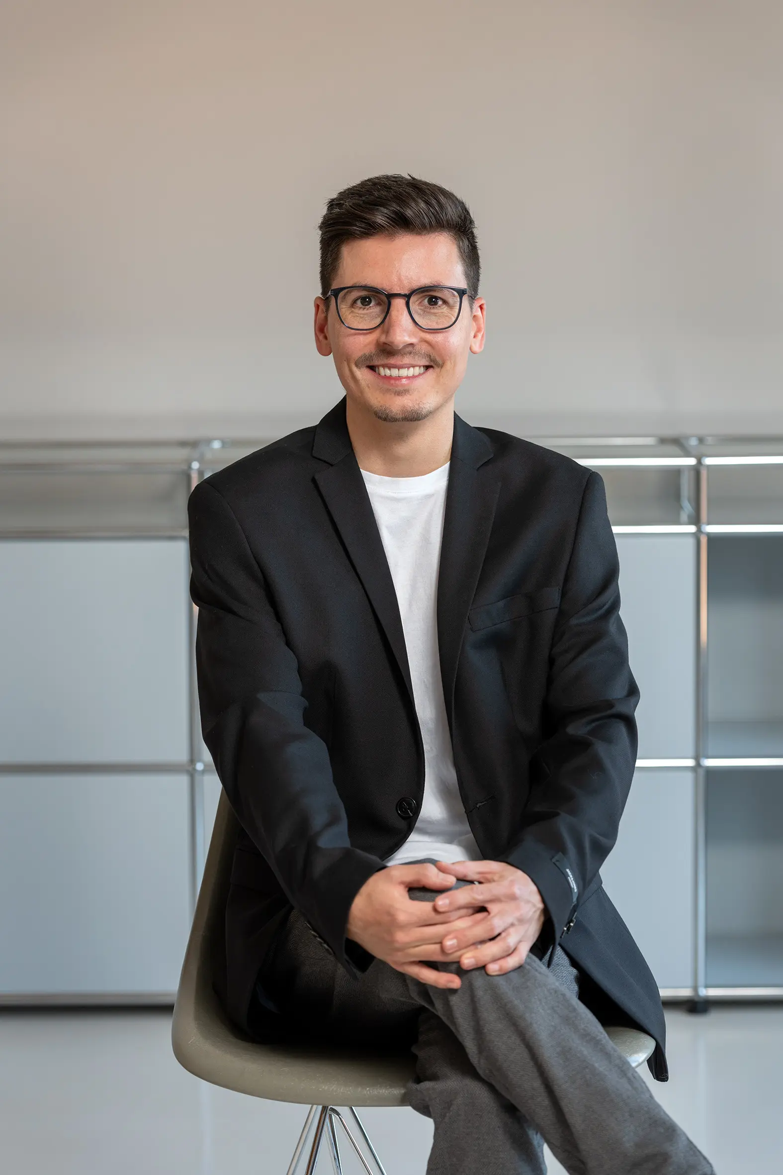 Porträt eines Mannes mit kurzen dunklen Haaren und Brille in einem schwarzen Blazer, lächelnd auf einem Stuhl, moderne Einrichtung im Hintergrund.